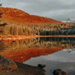 Tranquil mountain lake reflecting orange sunset-lit forest and snowy peaks in calm waters.