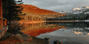 Tranquil mountain lake reflecting orange sunset-lit forest and snowy peaks in calm waters.