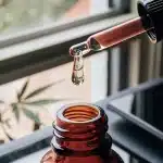 Huile de cannabis versée avec une pipette dans un flacon en verre ambré, sur une surface intérieure éclairée naturellement.