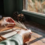 une main attache délicatement un petit bouquet d’herbes séchées près d’une fenêtre ensoleillée.