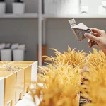 une main tient un petit sachet de graines au-dessus de plantes décoratives dans une boutique de jardinage.
